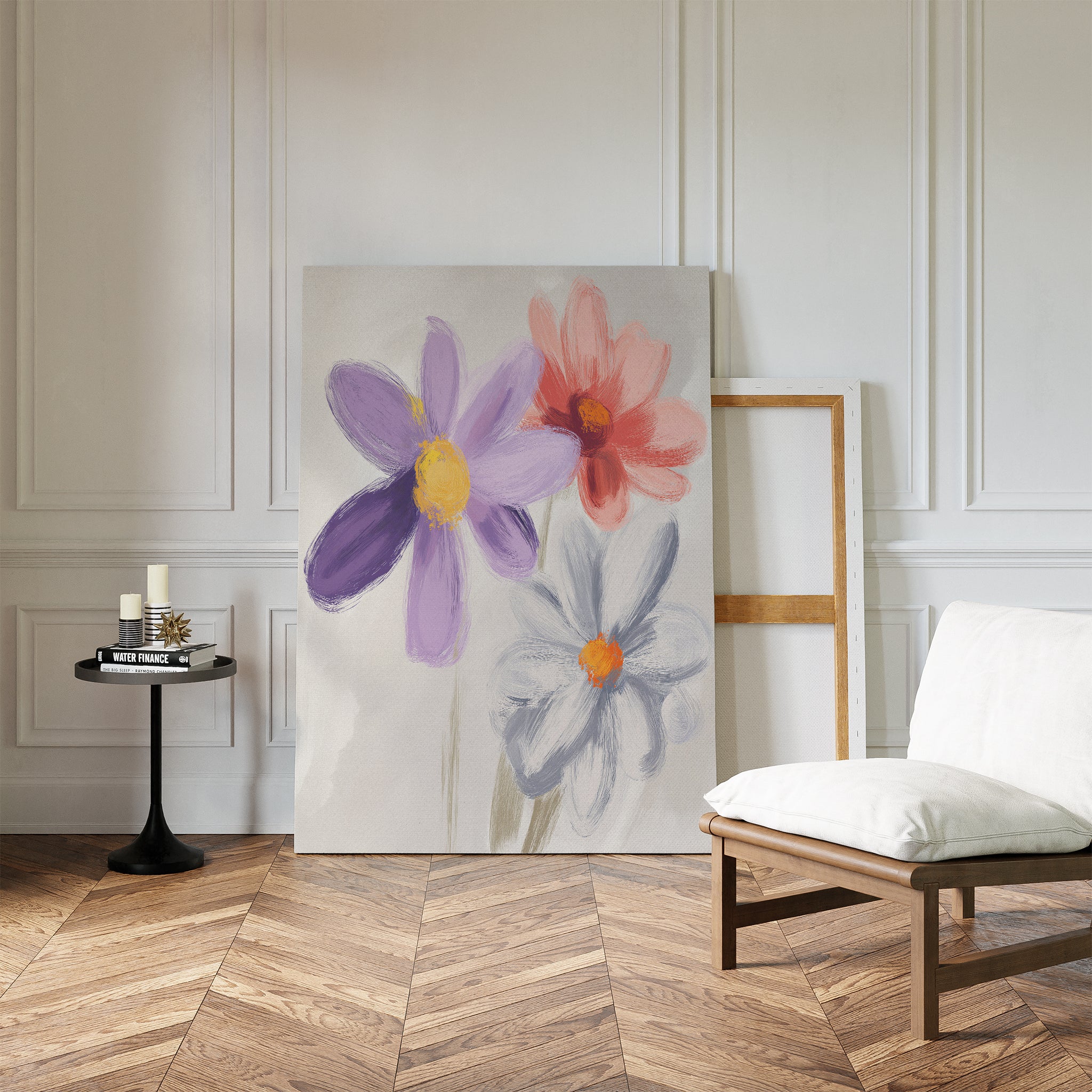 The Blooming Harmony Canvas Art - Vertical, featuring three painted flowers, leans against a white paneled wall in a stylish room with herringbone wood floors, a modern low chair, and a small black round table with candles and books.