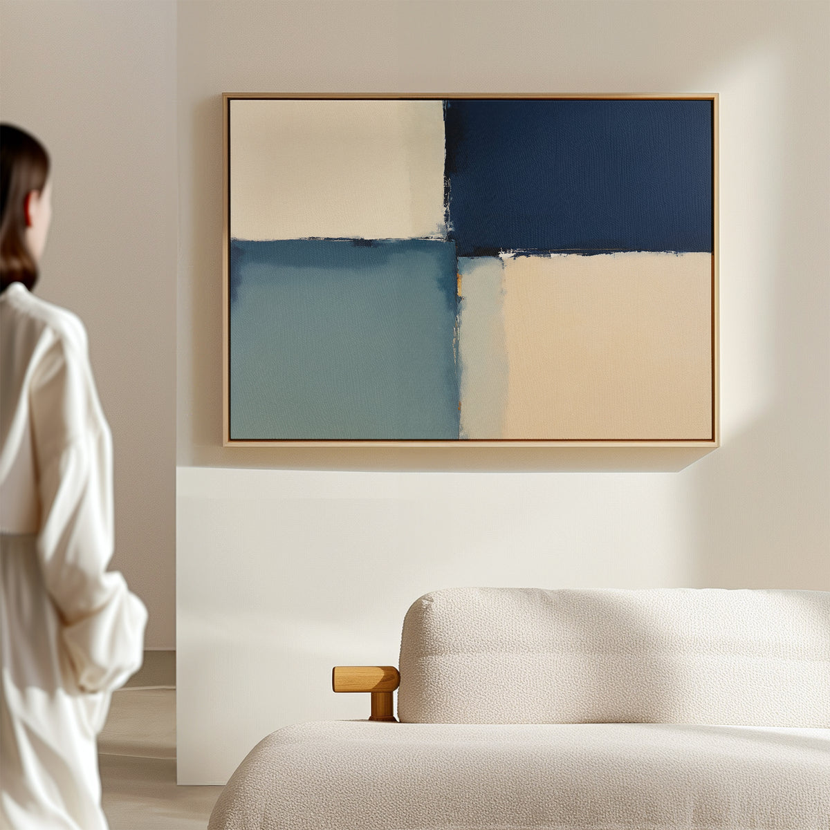 A person stands in a minimalistic room with a beige sofa, admiring the Blue Quarters Canvas Art - Horizontal, a contemporary abstract print in blue, cream, and tan that reflects the essence of modern living rooms.