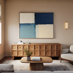 A modern living room with a light sofa, wooden coffee table, textured sideboard, and the Blue Quarters Canvas Art - Horizontal in blue and cream on the wall. A soft beige rug covers the floor, with a small vase of flowers on the table.