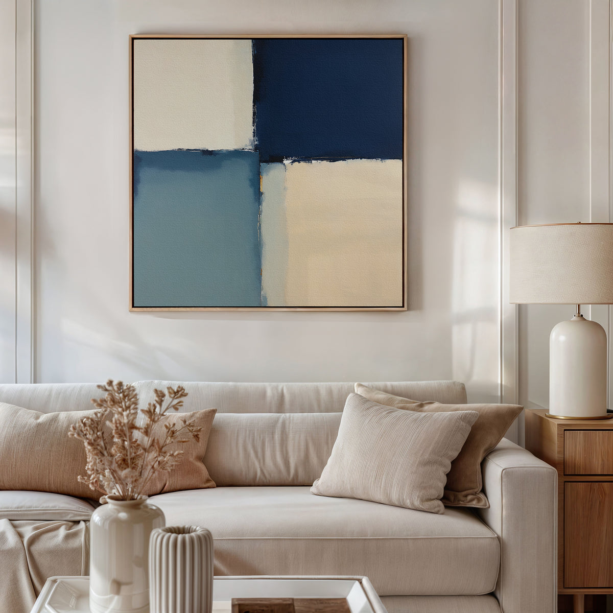 A modern living room with a beige sofa, neutral cushions, dried flowers in a vase, a table lamp, and the Blue Quarters Canvas Art - Square featuring navy, light blue, and cream geometric blocks above the sofa.