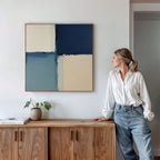A woman in a white blouse and jeans leans against a wooden cabinet, gazing at the Blue Quarters Canvas Art - Square, featuring blue and beige shapes. A plant and books on the cabinet create a modern living room vibe.
