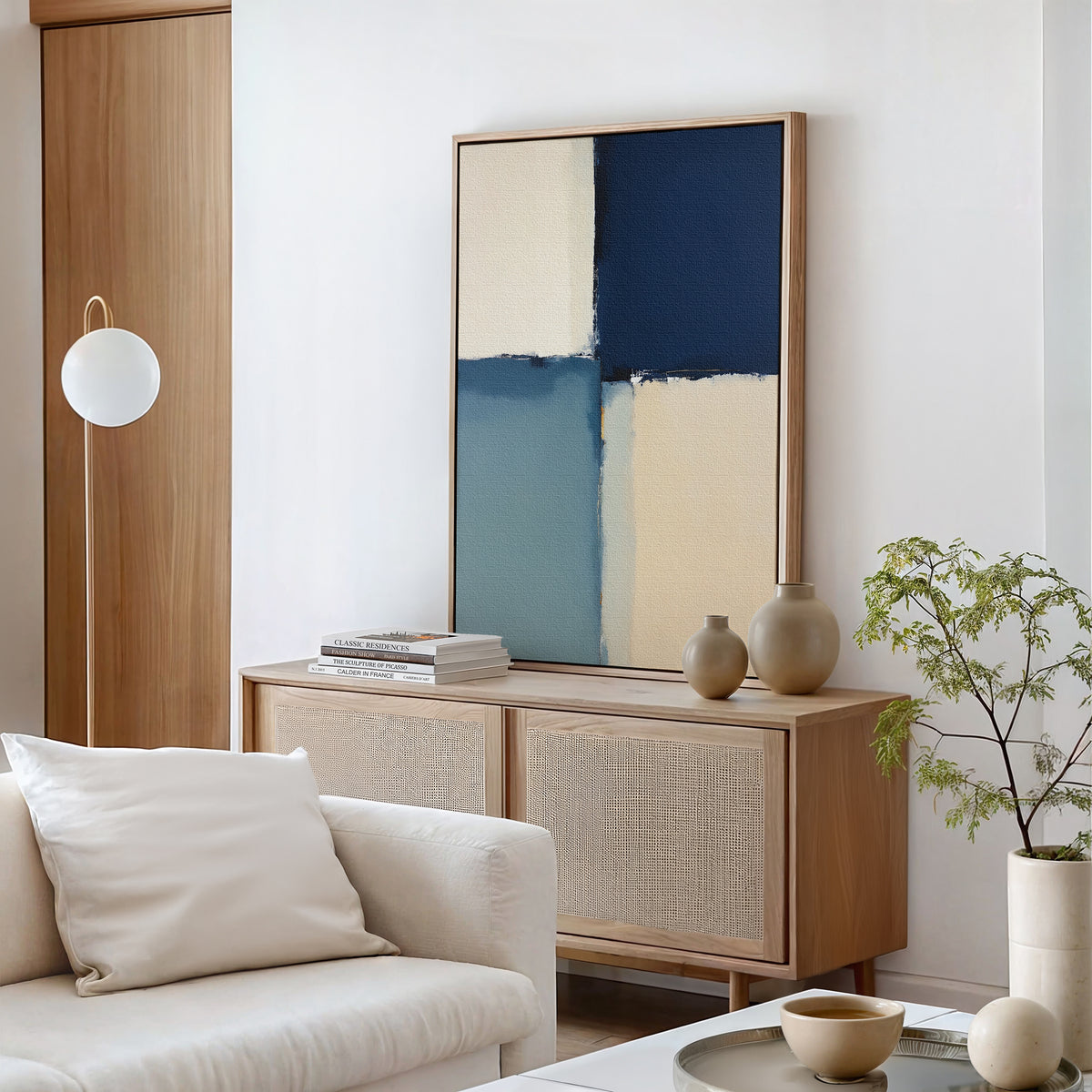 A modern living room with a light wood sideboard, Blue Quarters Canvas Art - Vertical in blue and beige tones, decorative vases, a small potted plant, and a white couch with a cushion. Minimalist decor in neutral colors.