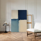 A modern room with herringbone wood floors displays the Blue Quarters Canvas Art - Vertical in blue and beige tones against a white-paneled wall, near a cushioned chair and a small round table with candles and books.