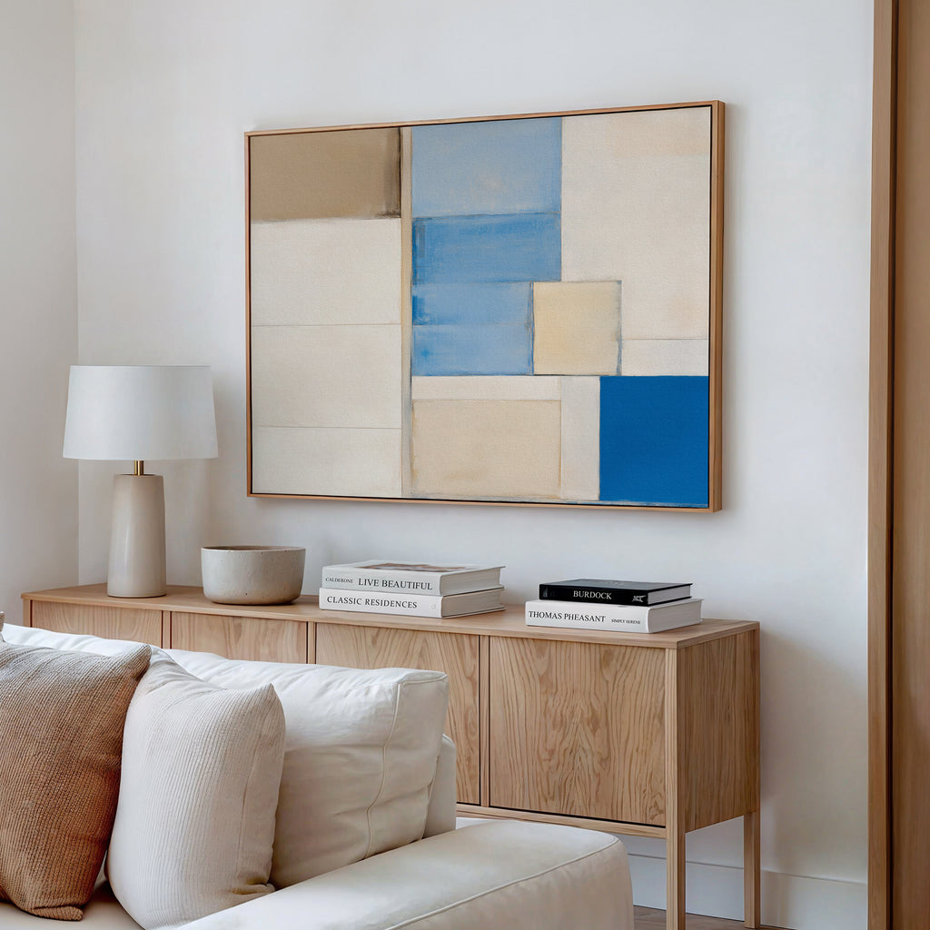 A modern living room features a wooden sideboard with books, a lamp, and a decor bowl. Above it hangs the Blue Tiles Composition Canvas Art - Horizontal in beige, blue, and white. A light sofa completes the contemporary decor.
