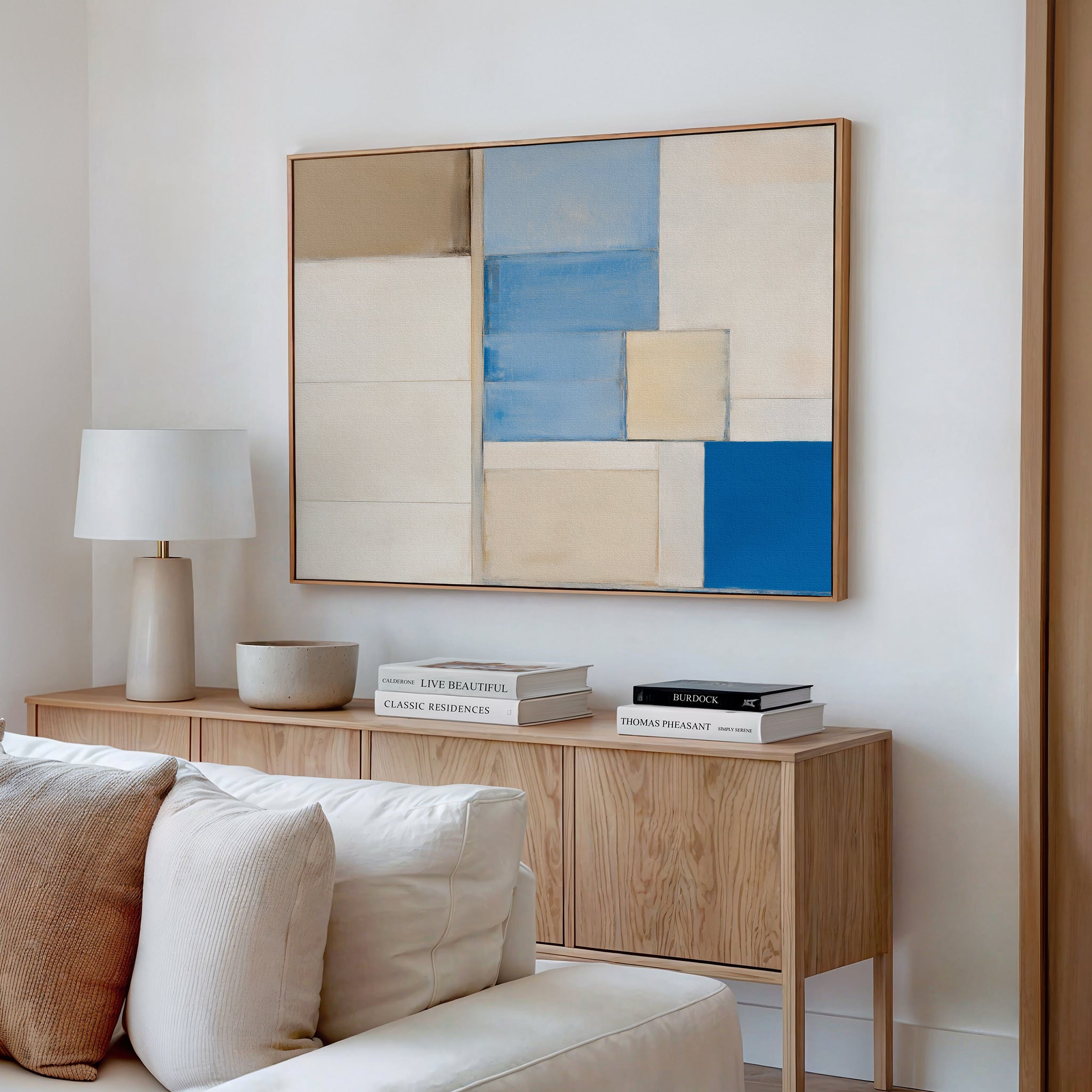A modern living room features a wooden sideboard with books, a lamp, and a decor bowl. Above it hangs the Blue Tiles Composition Canvas Art - Horizontal in beige, blue, and white. A light sofa completes the contemporary decor.