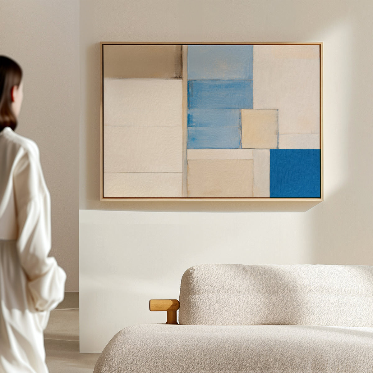 A woman in a white outfit stands in a bright, minimal room, admiring the Blue Tiles Composition Canvas Art—horizontal geometric wall decor in beige, white, and blue—displayed above a textured cream sofa for an elegant modern look.