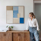 A woman in a white shirt and jeans leans against a wooden cabinet, admiring the Blue Tiles Composition Canvas Art - Square, featuring blue, beige, and white rectangles. Books and a small plant accent the modern living room decor.