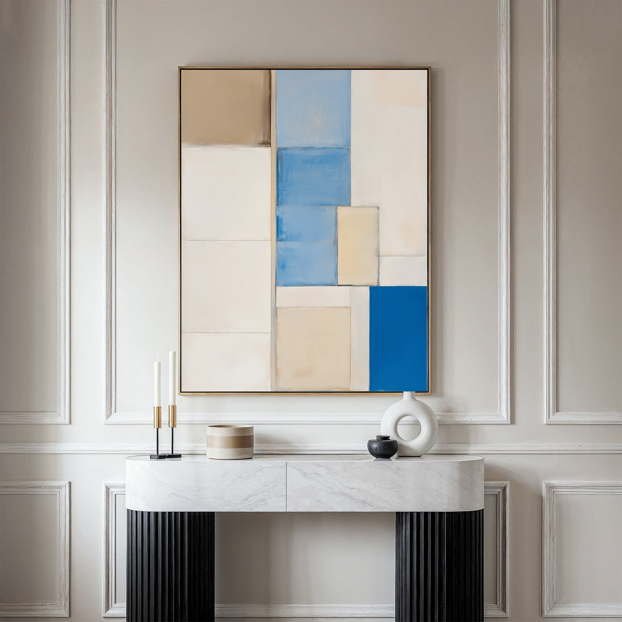 A minimalist console table with decorative objects stands beneath the Blue Tiles Composition Canvas Art - Vertical, which features beige, cream, blue, and tan geometric shapes against elegant paneled walls.