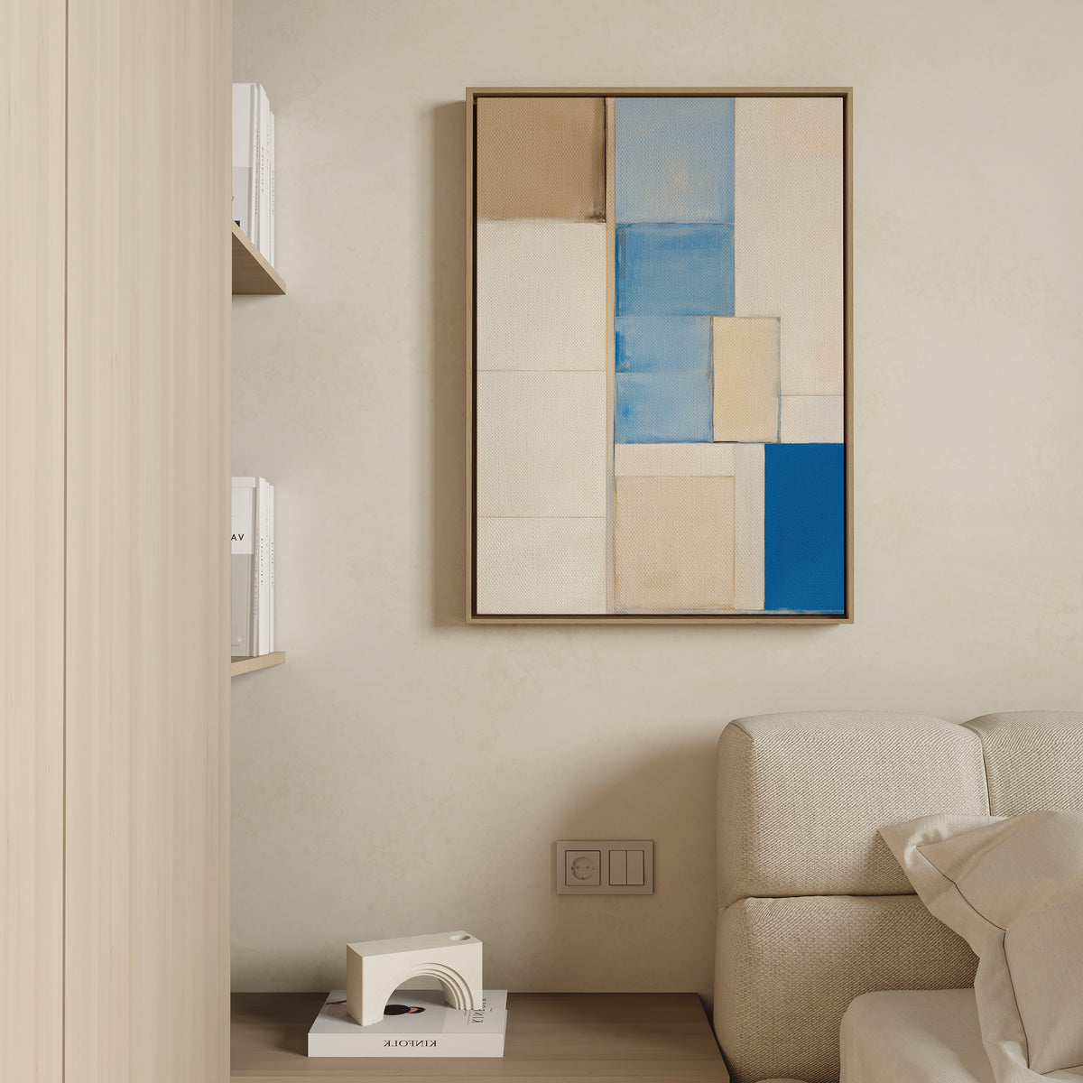 A minimal living room corner with a beige couch, a small wooden shelf of books, and the Blue Tiles Composition Canvas Art - Vertical in beige, brown, and blue hanging on a cream wall. A decorative object rests on the floor beside the sofa.