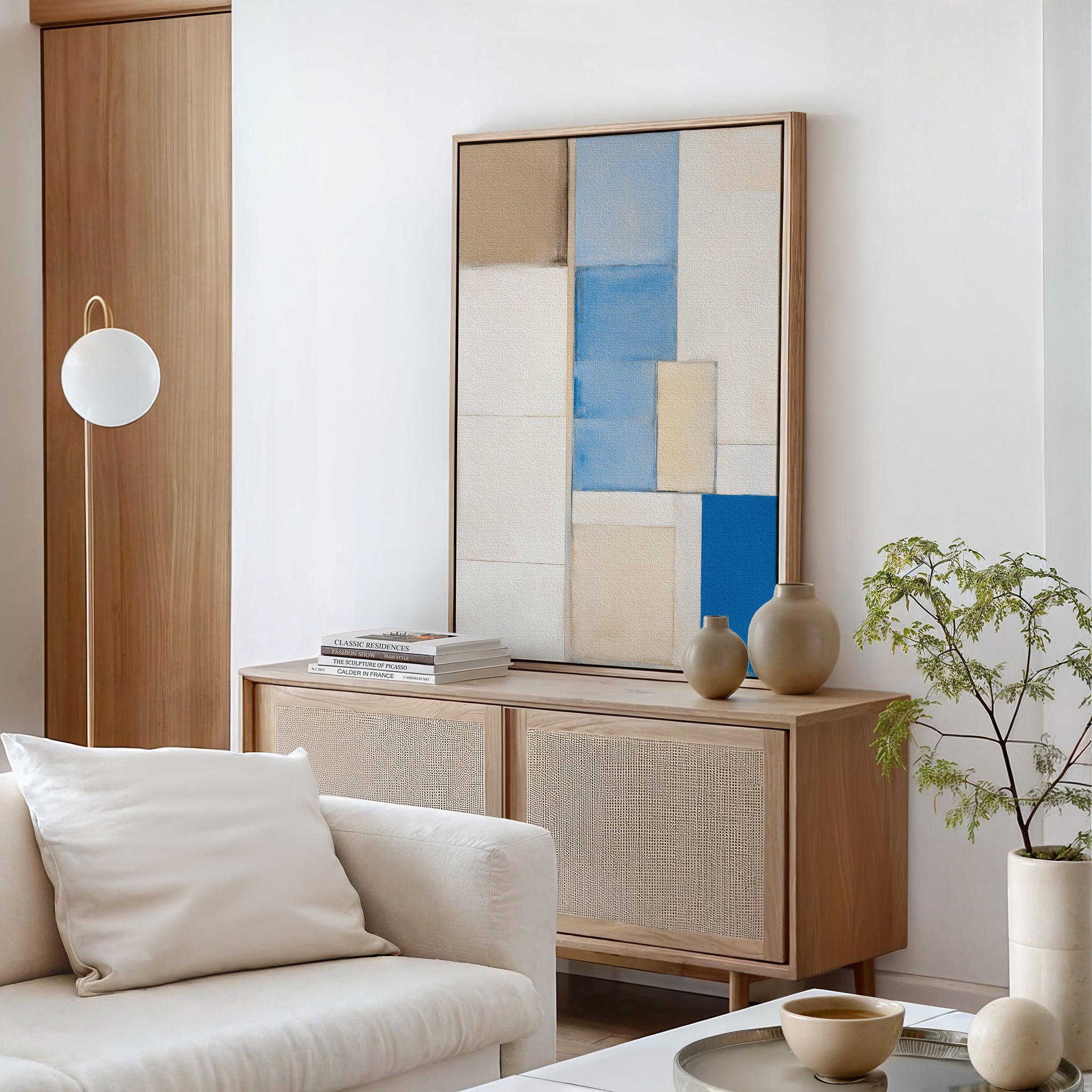Minimalist living room featuring a light wood sideboard, the Blue Tiles Composition Canvas Art - Vertical in beige and blue, ceramic vases, a potted plant, round lamp, white sofa, and a stack of books on the cabinet.