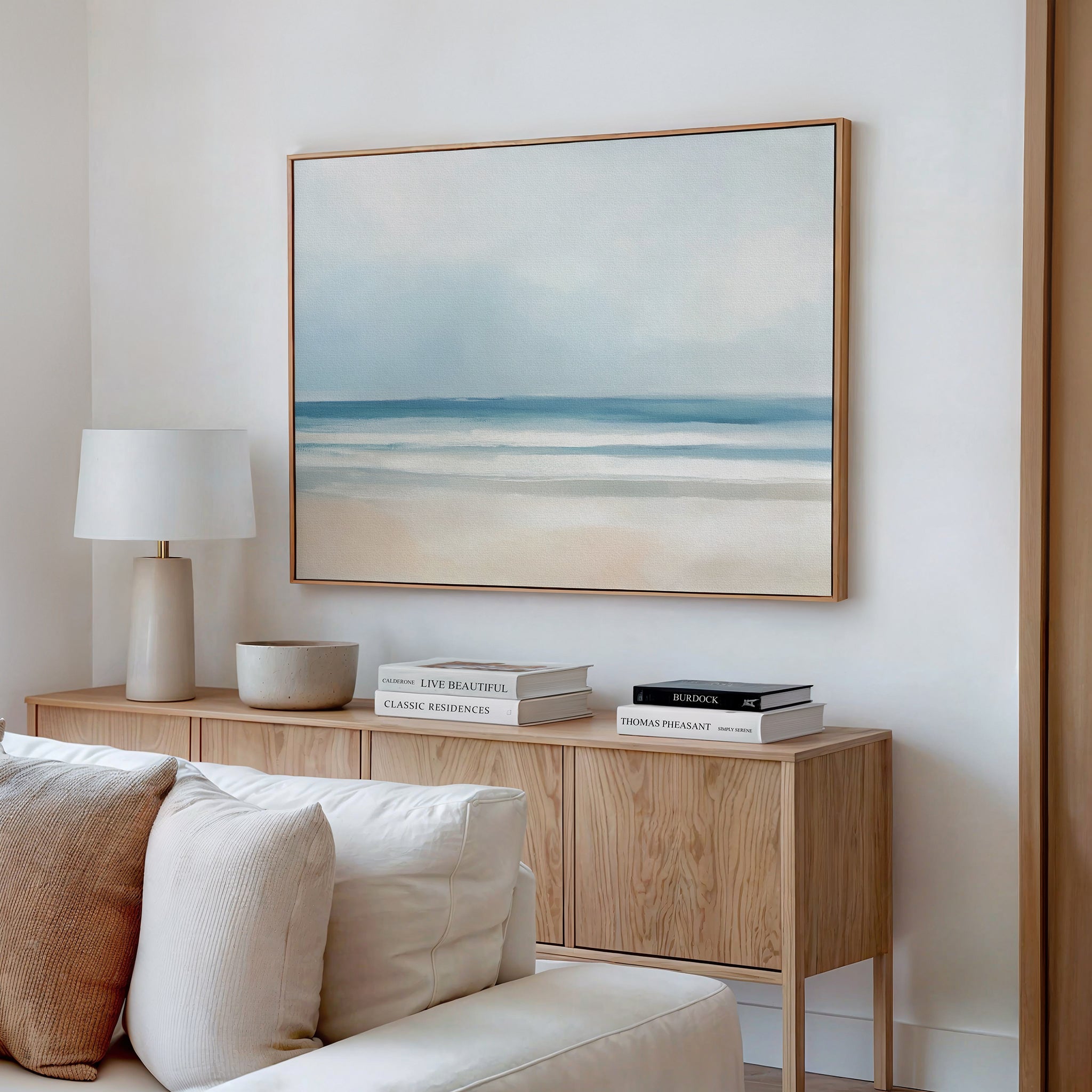 A minimalist living room with a beige couch, wooden sideboard, books, and a white lamp features the Blurred Beach Scene Canvas Art - Horizontal as tranquil wall decor. The overall look is light and neutral.