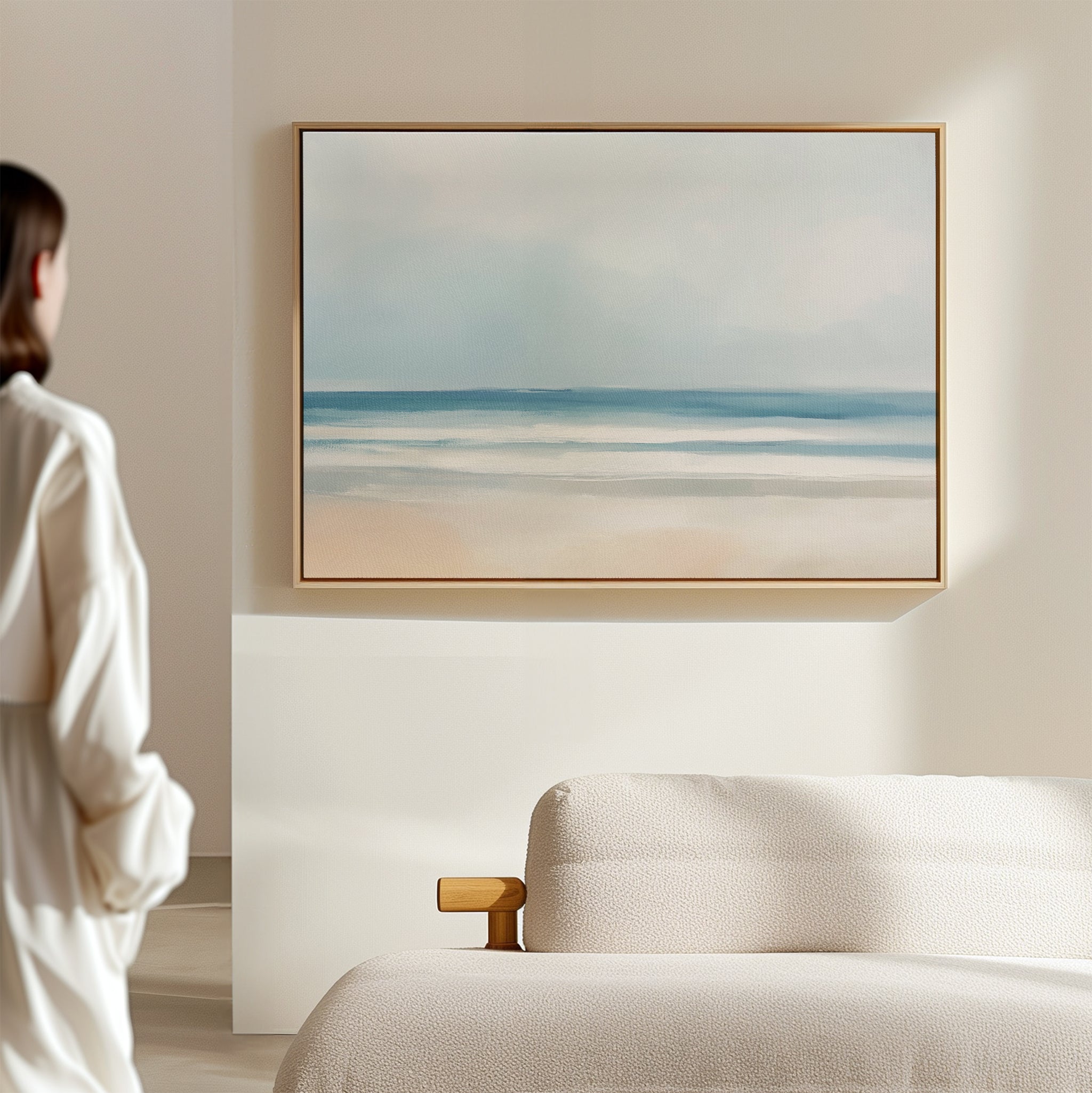 In a minimalist, light-filled room, someone admires the Blurred Beach Scene Canvas Art - Horizontal, a serene seascape in soft blue and beige tones, elegantly framed and displayed above a textured white sofa.
