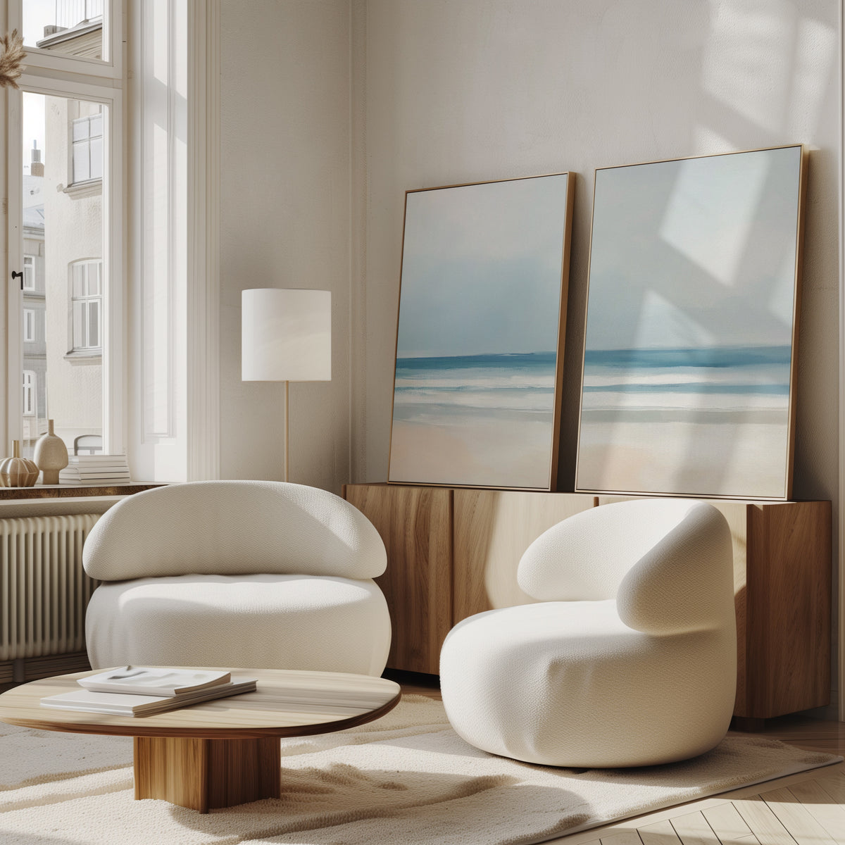 A bright, modern living room with two white lounge chairs, a low wooden coffee table, and serene decor featuring the Blurred Beach Scene - Set Of 2 wall art on a wooden sideboard. Sunlight streams in from large windows.