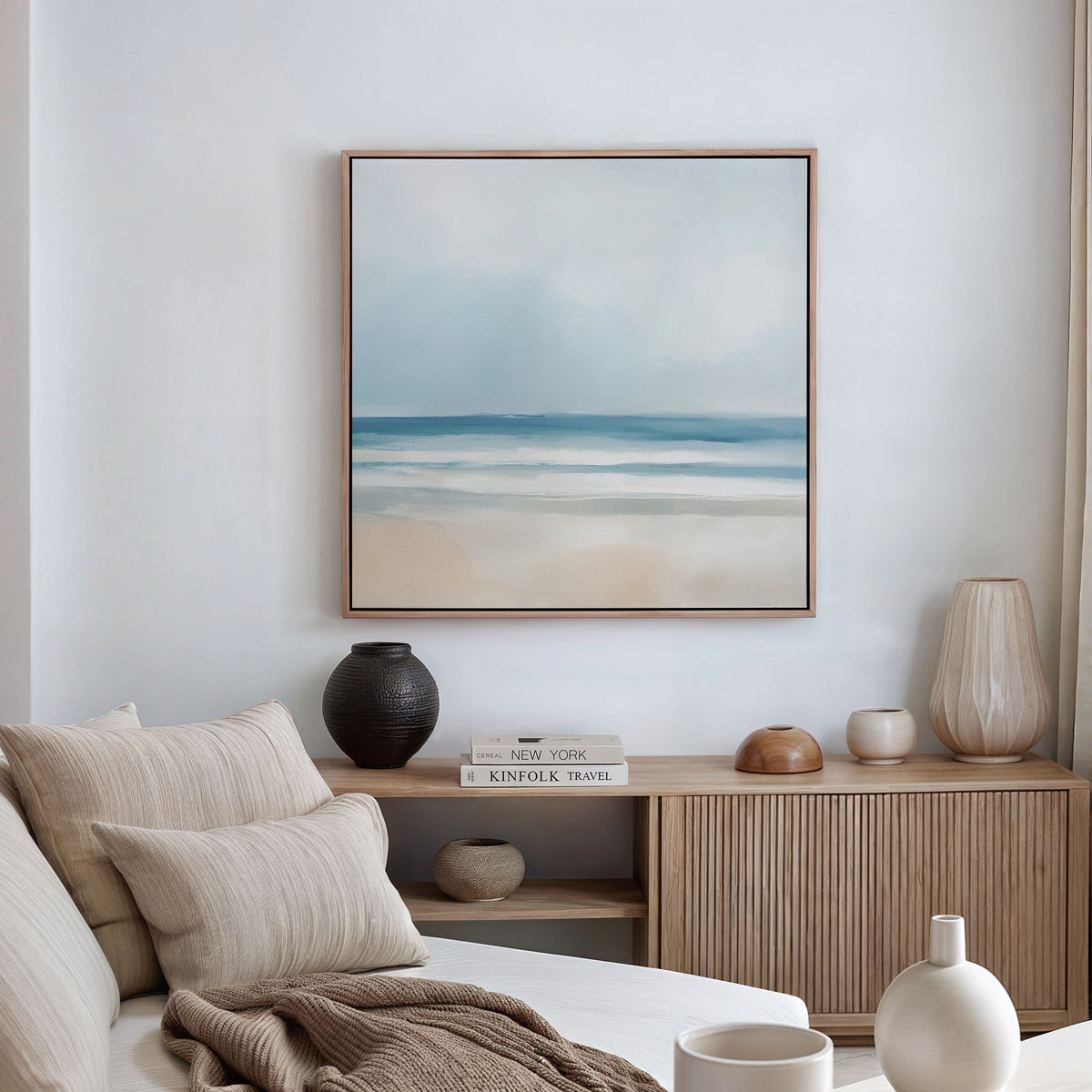 A minimal, modern living room with a beige sofa, neutral cushions, wooden sideboard, decorative vases, stacked books, and the Blurred Beach Scene Canvas Art - Square displayed on a white wall. Soft natural light fills the space.