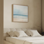A minimal, cozy bedroom features a cream upholstered headboard, white bedding, and the Blurred Beach Scene Canvas Art - Square in soft blue, white, and beige above the bed. A bare branch adds decor to the nightstand.