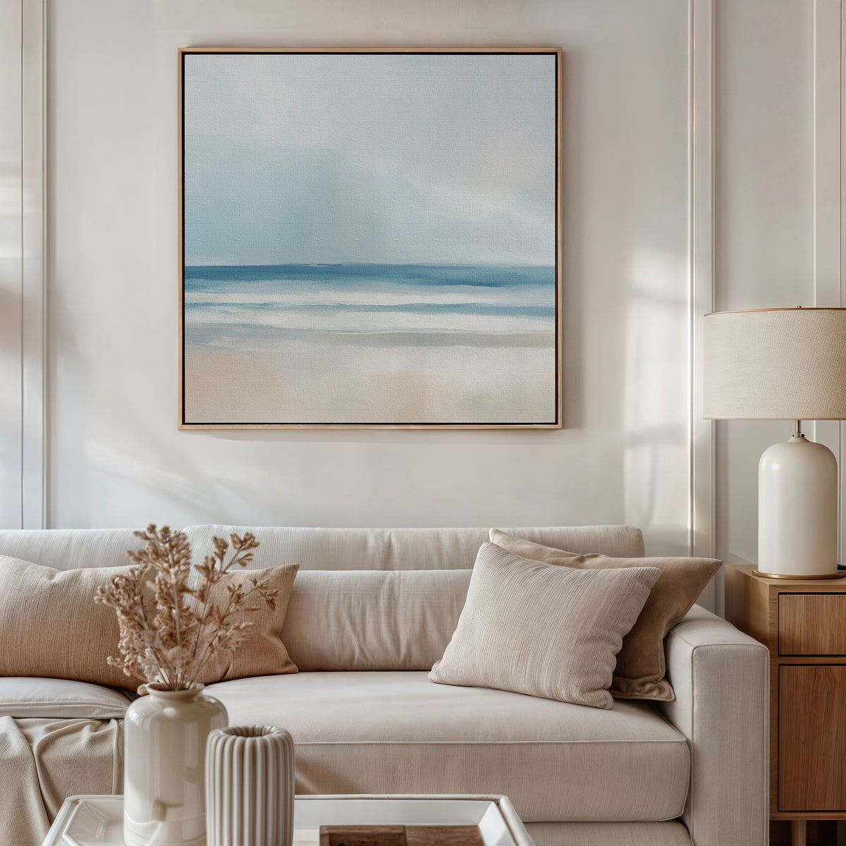 A neutral-toned living room features a beige sofa with light cushions, a side table holding a ceramic lamp, and the Blurred Beach Scene Canvas Art - Square above the couch. Dried flowers in a vase sit on the coffee table.