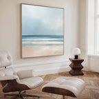 A modern, minimalist room featuring a white leather lounge chair and ottoman, a wooden side table with a white lamp, and tranquil decor—a Blurred Beach Scene Canvas Art - Square displaying a serene coastal view in soft blue and beige tones.