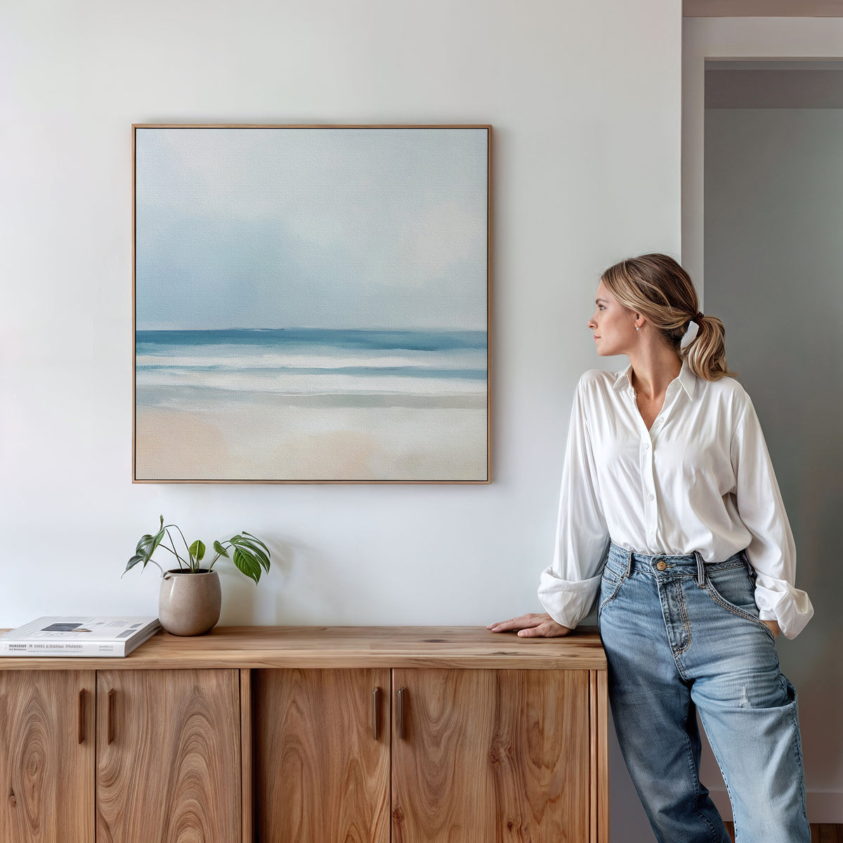 A woman in a white blouse and jeans leans on a wooden cabinet, gazing at the Blurred Beach Scene Canvas Art - Square. A potted plant and a closed book rest neatly on the cabinet.
