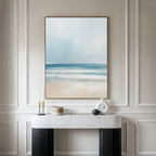 A minimalist console table with decor stands against a paneled wall, topped by the Blurred Beach Scene Canvas Art - Vertical, an abstract coastal print in soft blue, white, and beige—ideal for serene beach-inspired decor.