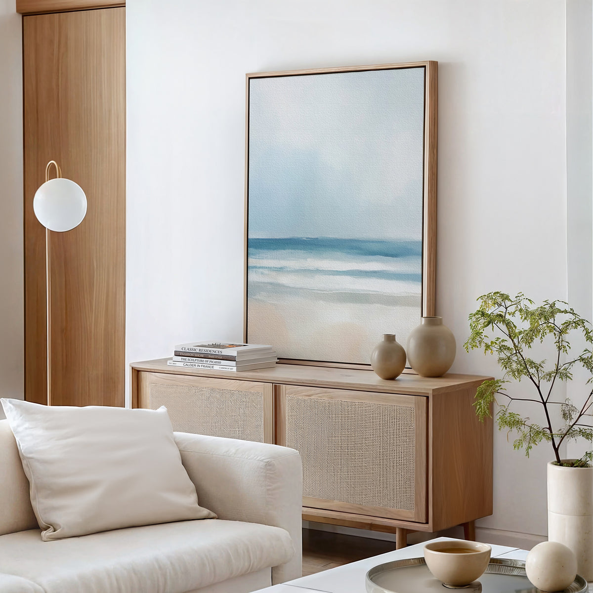 A modern, minimalist living room with neutral tones features a couch with a white pillow, wooden sideboard, ceramic vases, potted plant, and wall art—a Blurred Beach Scene Canvas Art - Vertical centerpiece.