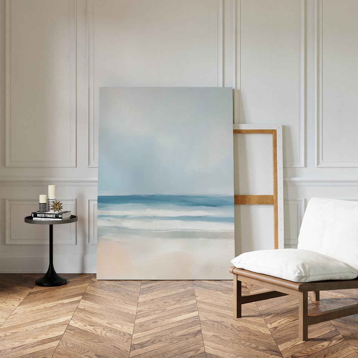 A minimalist room with wood herringbone floors, white paneled walls, a cushioned chair, a small round table holding candles and books, and serene decor featuring the Blurred Beach Scene Canvas Art - Vertical leaning against the wall.