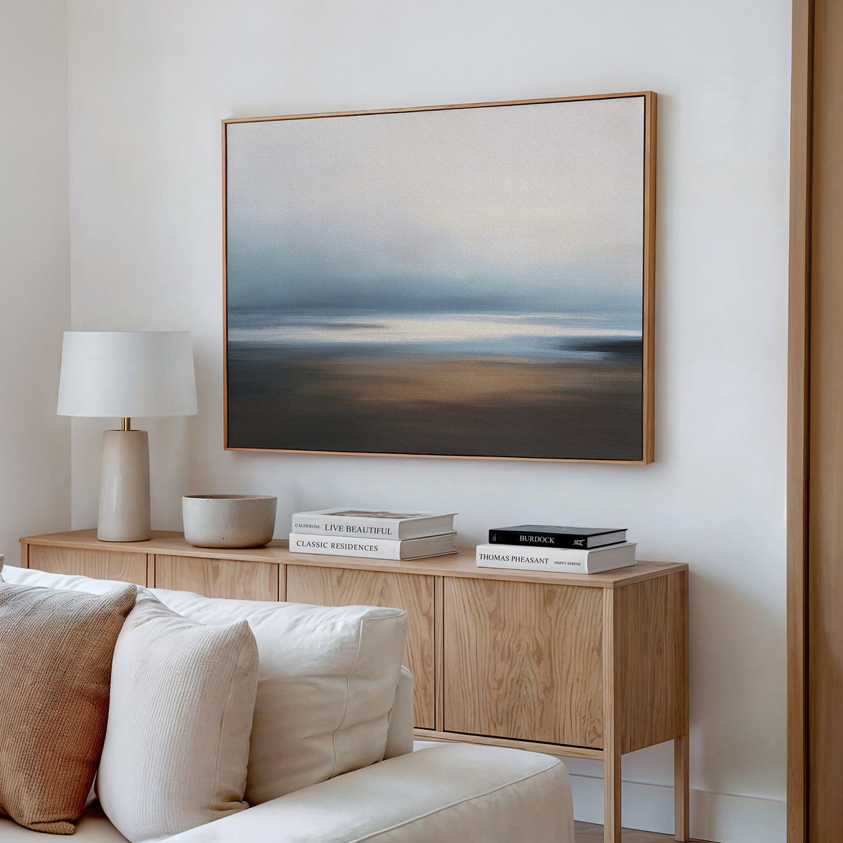 A modern living room with a light wood sideboard, books, lamp, and bowl showcases the Blurred Seascape Canvas Art - Horizontal above a cream sofa—its soft blue, beige, and gray hues are ideal for lovers of coastal serenity decor.