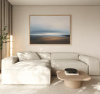 A modern living room with a light beige sectional, round wooden coffee table with a black bowl, tall plant, and the Blurred Seascape Canvas Art - Horizontal above the sofa for a touch of coastal serenity.
