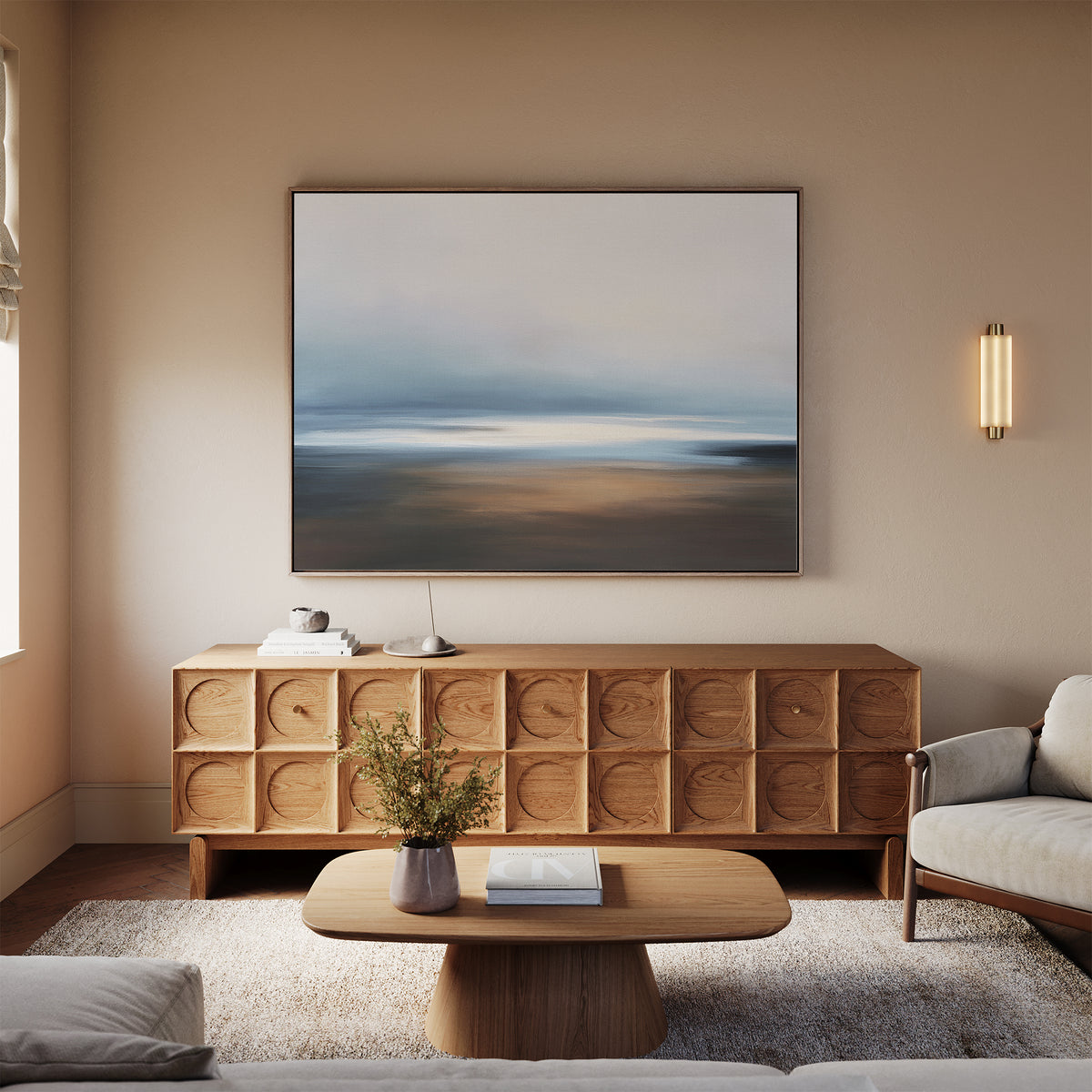 A modern living room featuring the Blurred Seascape Canvas Art - Horizontal above a wooden cabinet, with a light sofa, small coffee table, neutral walls, and wall-mounted lighting for serene coastal décor.