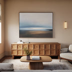A modern living room featuring the Blurred Seascape Canvas Art - Horizontal above a wooden cabinet, with a light sofa, small coffee table, neutral walls, and wall-mounted lighting for serene coastal décor.