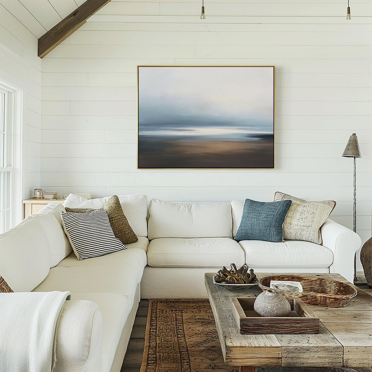 A cozy, bright living room features a white sectional sofa, assorted throw pillows, and a rustic wooden coffee table. The space exudes coastal serenity with the Blurred Seascape Canvas Art - Horizontal displayed above the couch on a shiplap wall.