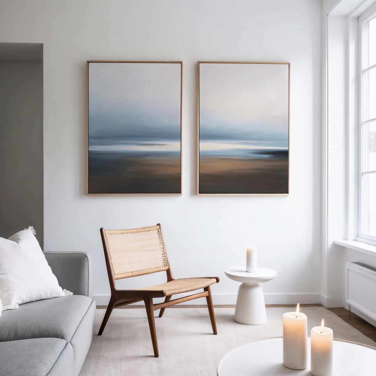 A bright, minimalist living room with a gray sofa, wooden chair, and white side table features the Blurred Seascape - Set Of 2 wall art prints in calming blue and brown tones, enhancing the coastal serenity decor.