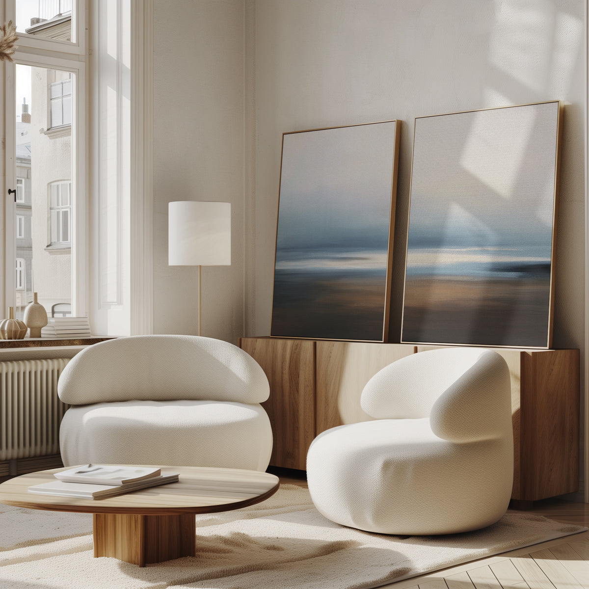 A modern living room features two curved white armchairs, a low wooden coffee table, a floor lamp, and the Blurred Seascape - Set Of 2 wall art above the sideboard, creating coastal serenity with neutral tones and soft natural light.