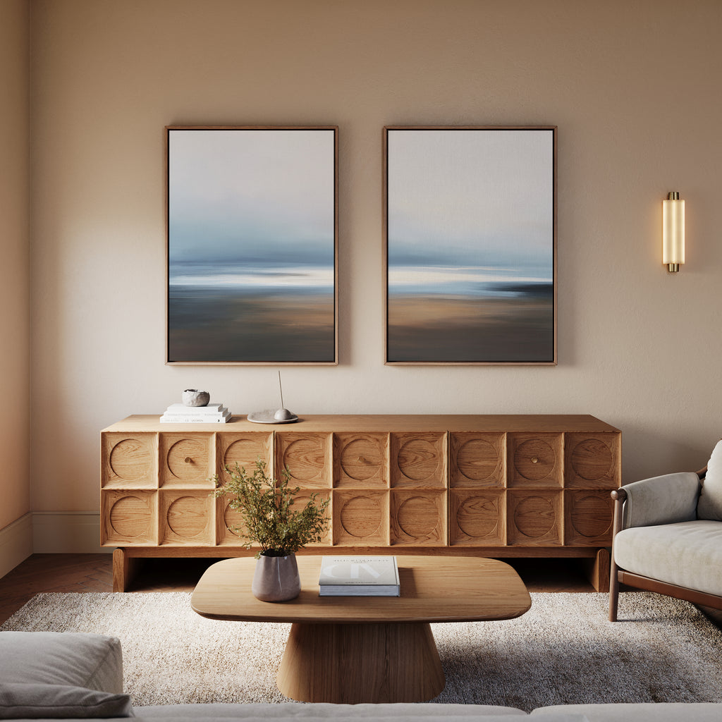 A modern living room with light walls, a wooden sideboard with geometric patterns, a wooden coffee table, a light sofa, a plant, and the "Blurred Seascape - Set Of 2" wall art above the sideboard for coastal serenity.