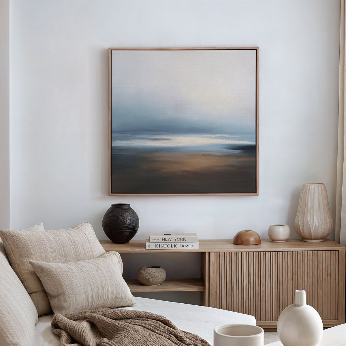 A cozy, modern living room features a beige sofa with neutral pillows, a wooden sideboard with vases and books, and the Blurred Seascape Canvas Art - Square as atmospheric wall art on the crisp white wall.