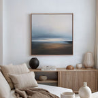 A cozy, modern living room features a beige sofa with neutral pillows, a wooden sideboard with vases and books, and the Blurred Seascape Canvas Art - Square as atmospheric wall art on the crisp white wall.