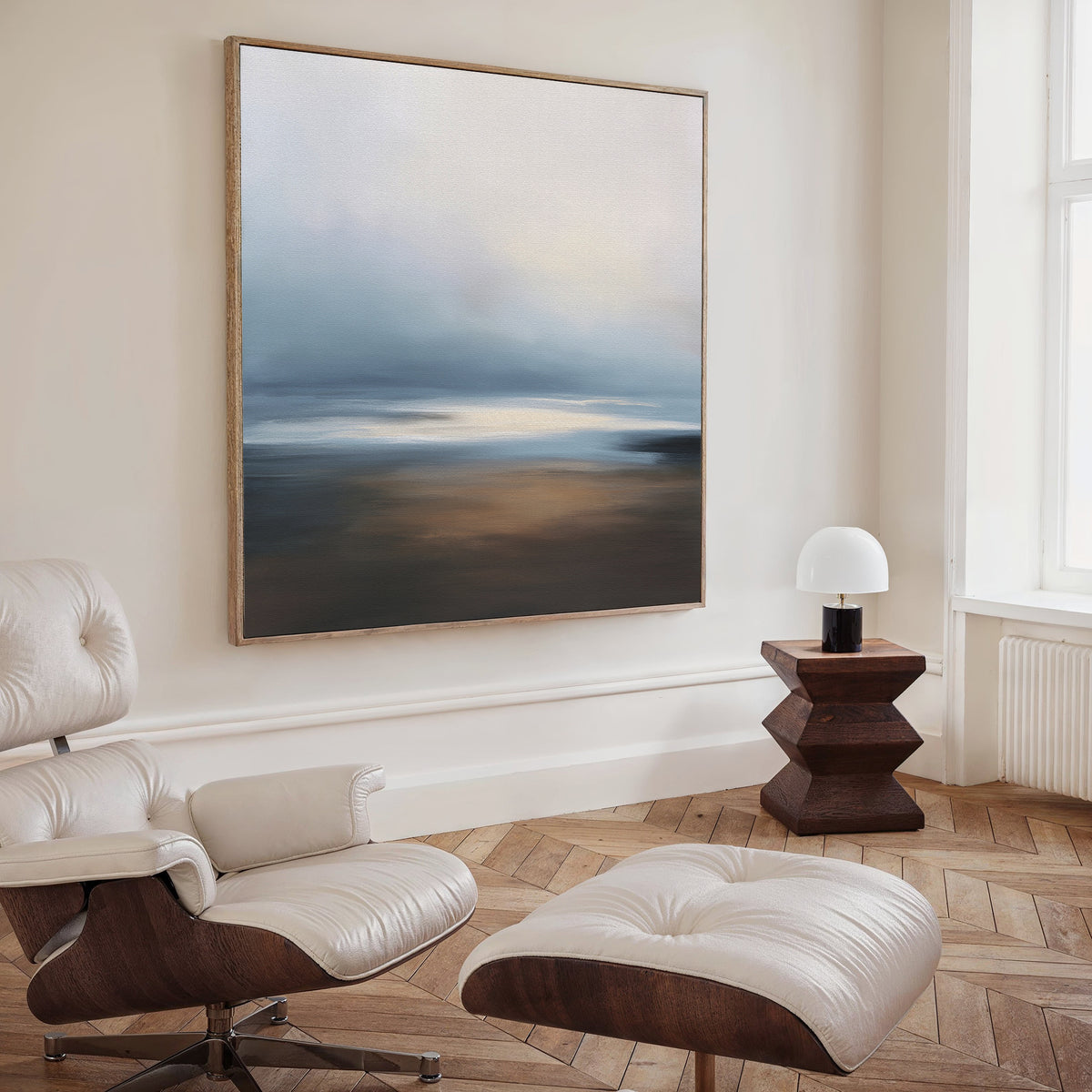 A modern, minimalist living space featuring a white leather lounge chair and ottoman, a geometric wooden side table with a small lamp, and the Blurred Seascape Canvas Art - Square adding an abstract touch to the wall.