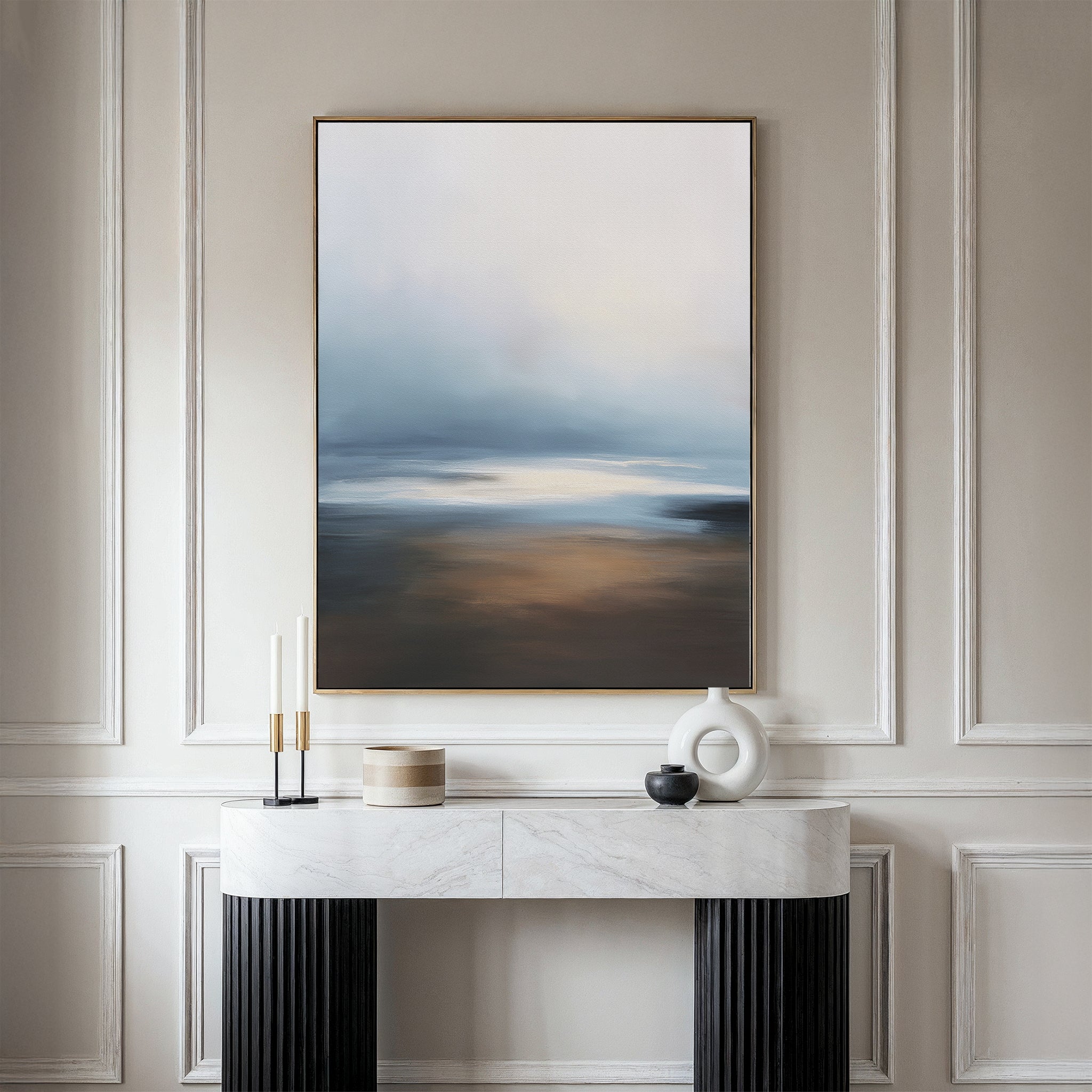 A minimalist console table with decorative objects sits against a paneled wall, topped by the Blurred Seascape Canvas Art - Vertical, featuring soft blue, white, and brown tones for an elegant, modern touch of coastal serenity.
