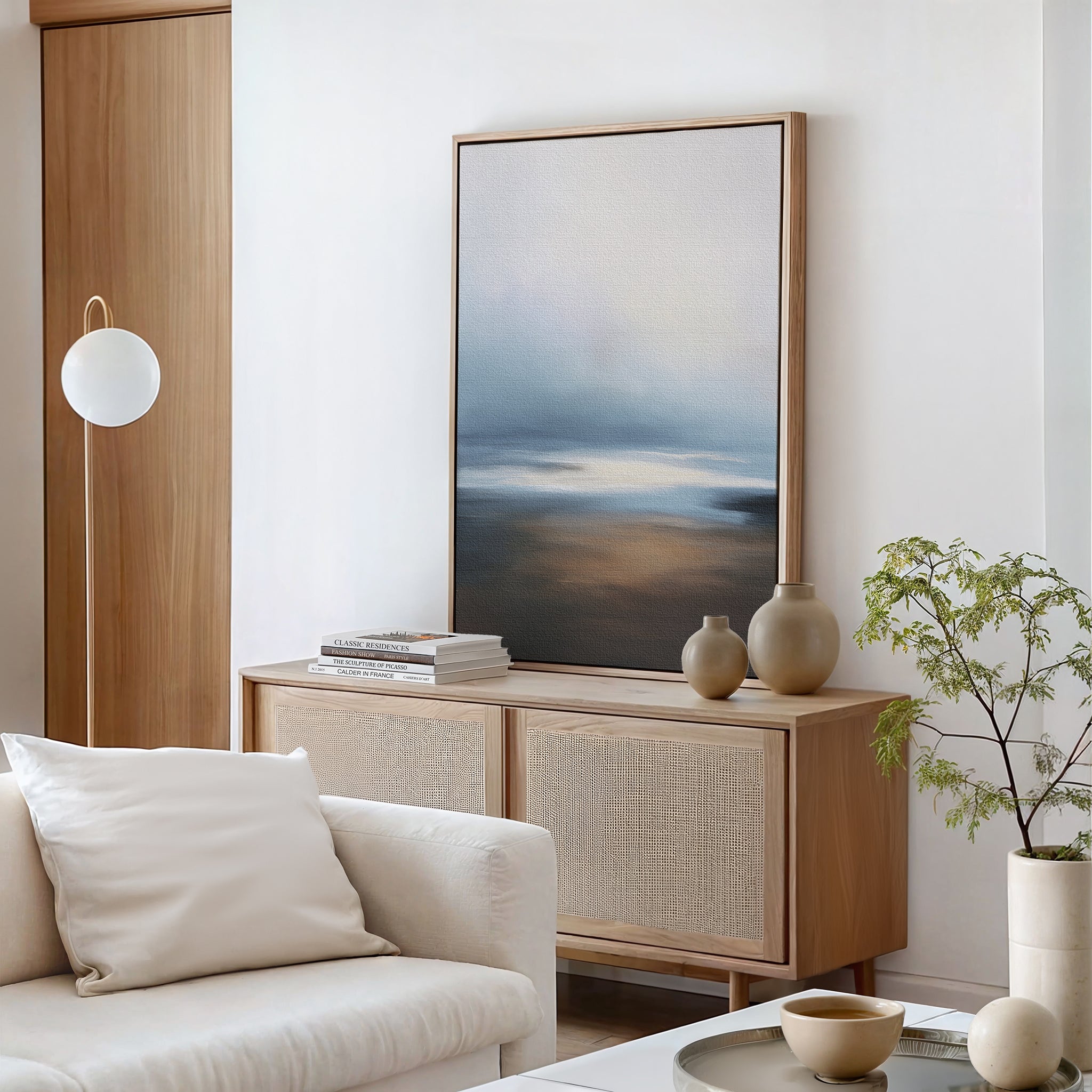 A minimalist living room featuring a light wood sideboard, the Blurred Seascape Canvas Art - Vertical for coastal vibes, neutral vases, stacked books, a small potted plant, white sofa with cushions, and a round hanging light.