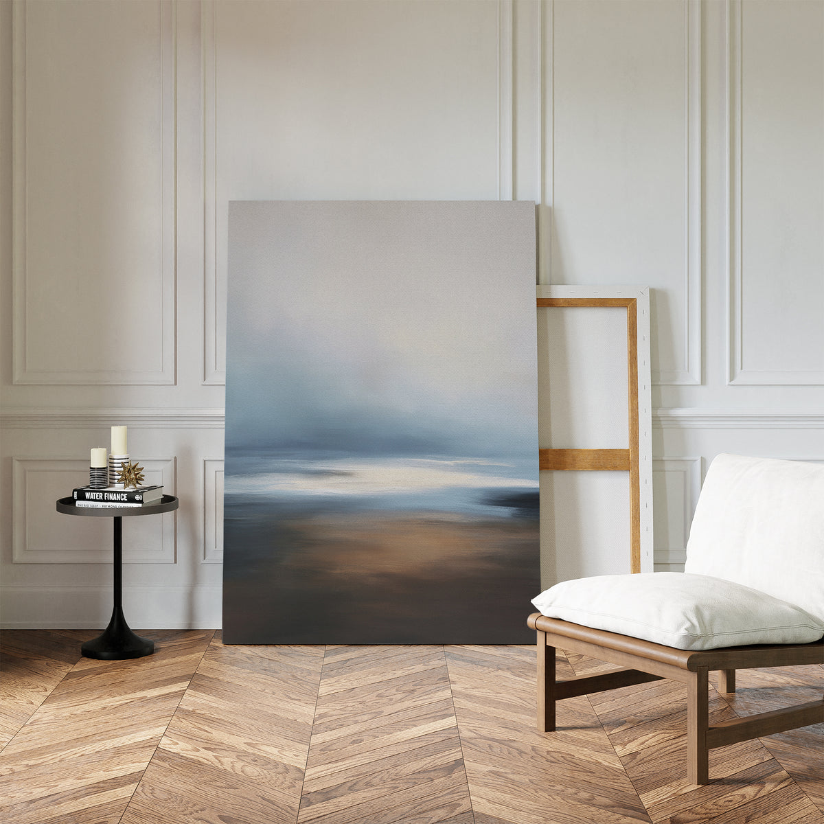 The Blurred Seascape Canvas Art - Vertical features soft blue, gray, and brown tones and leans against a white-paneled wall, adding coastal serenity to a room with herringbone floors, a cushioned chair, and a small black side table with candles.