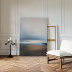 The Blurred Seascape Canvas Art - Vertical features soft blue, gray, and brown tones and leans against a white-paneled wall, adding coastal serenity to a room with herringbone floors, a cushioned chair, and a small black side table with candles.