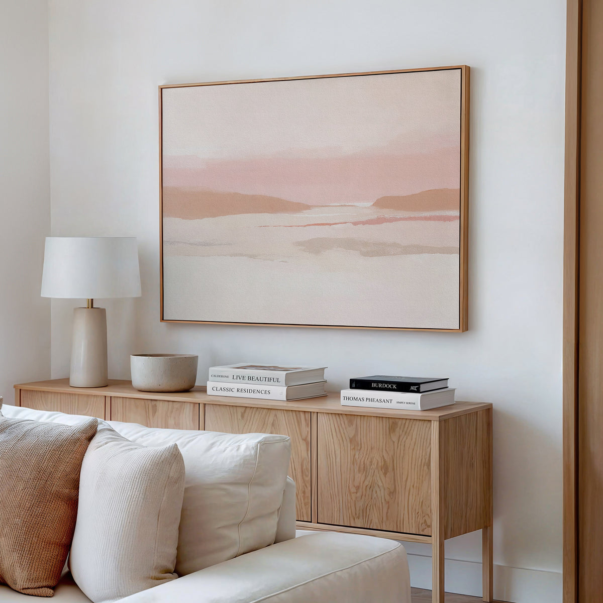 A minimalist living room features a wooden sideboard, white lamp, books, and bowl. Above the cream sofa with textured pillows hangs the Blush Mesa Canvas Art - Horizontal, adding earth tones and tranquility to the space.