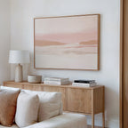 A minimalist living room features a wooden sideboard, white lamp, books, and bowl. Above the cream sofa with textured pillows hangs the Blush Mesa Canvas Art - Horizontal, adding earth tones and tranquility to the space.