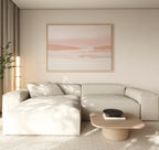 A modern living room with a light beige sectional sofa, wooden coffee table with a black bowl, tall plant, and the Blush Mesa Canvas Art - Horizontal—a serene landscape in soft pink and beige—completes the earth tone decor.