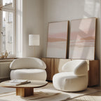 A modern living room with the Blush Mesa - Set Of 2 white rounded armchairs, a low wooden coffee table, a floor lamp, and minimalist pink and beige wall art on a wooden cabinet. Sunlight streams in through large windows.