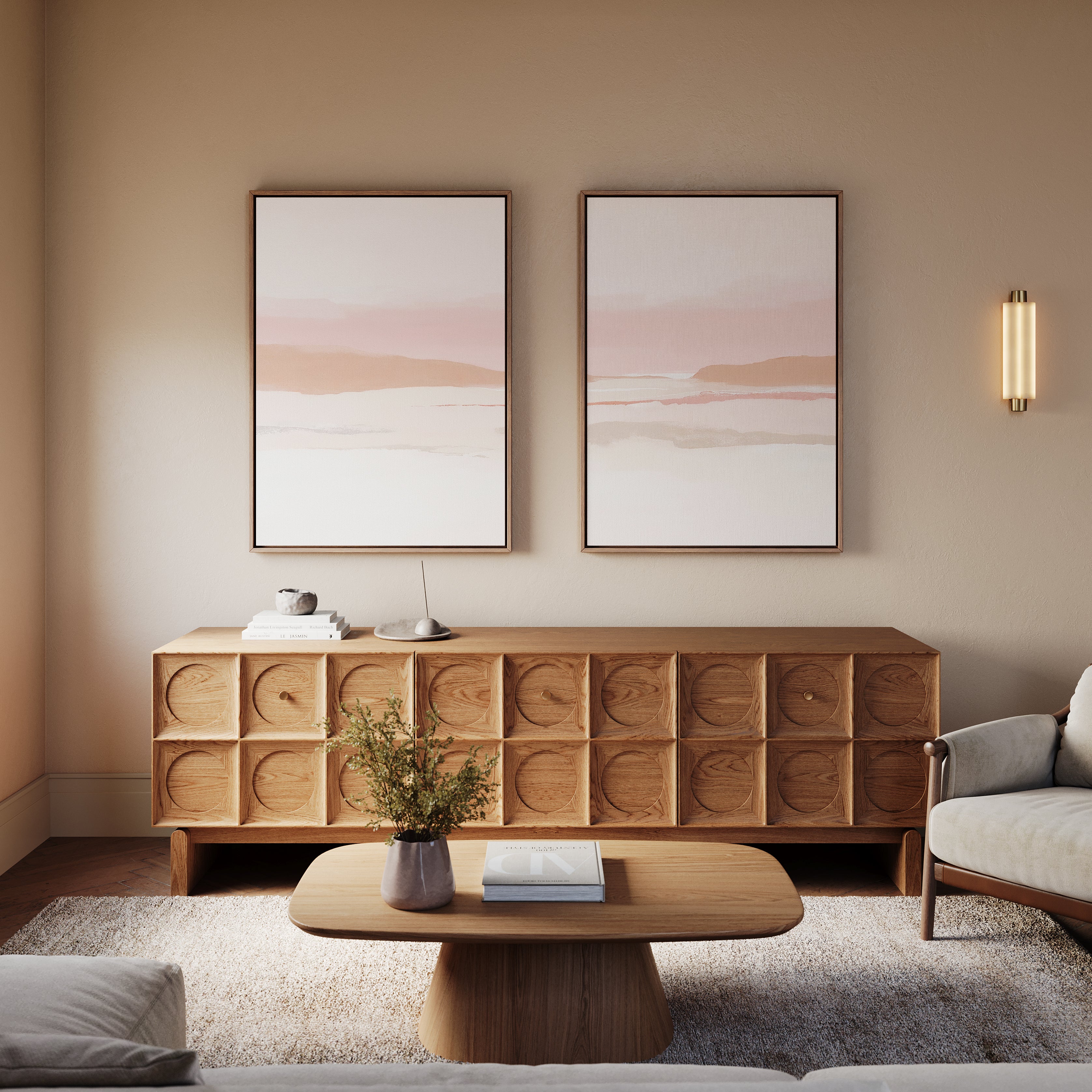 A minimalist living room with light walls and earth tones features the Blush Mesa - Set Of 2 abstract art above a wooden sideboard, plus a matching coffee table, small plant, and a cozy armchair in soft, neutral shades.