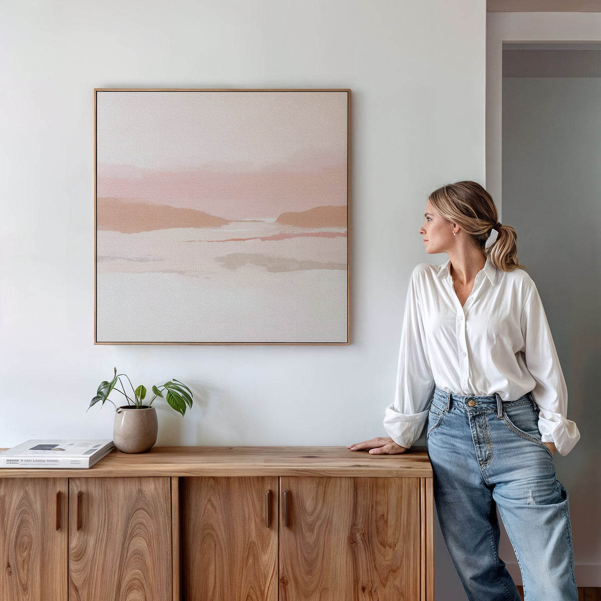 A woman in a white blouse and jeans leans against a wooden cabinet, gazing at the Blush Mesa Canvas Art - Square in soft earth tones. A small plant and a closed book sit on the cabinet.