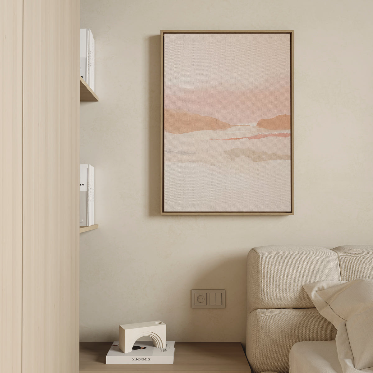 Minimalist living space with a beige couch, wooden side table, shelves of books, and the Blush Mesa Canvas Art - Vertical—a soft pink and earth-toned abstract landscape—displayed on a cream-colored wall.