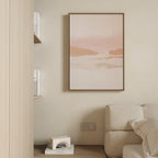 Minimalist living space with a beige couch, wooden side table, shelves of books, and the Blush Mesa Canvas Art - Vertical—a soft pink and earth-toned abstract landscape—displayed on a cream-colored wall.