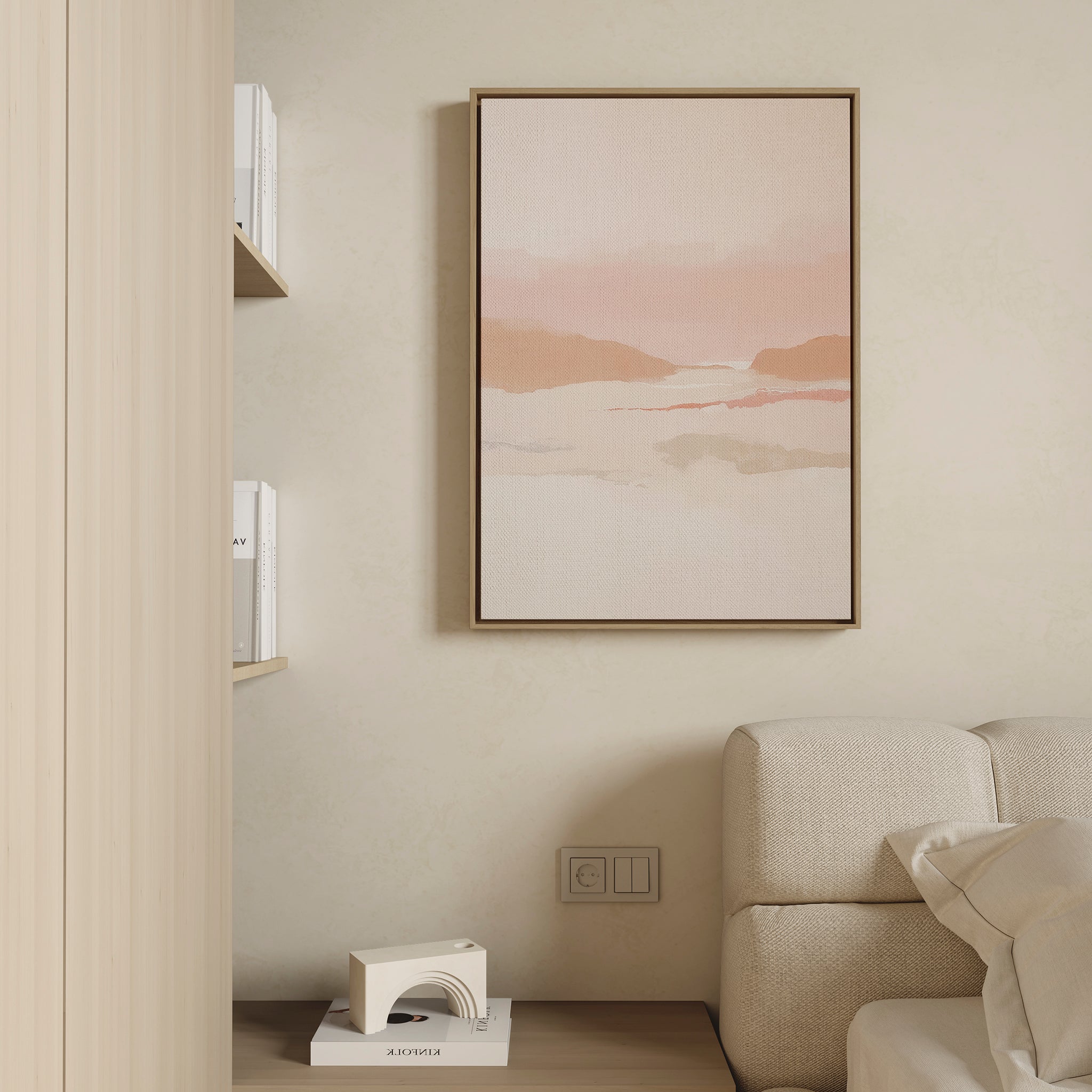 Minimalist living space with a beige couch, wooden side table, shelves of books, and the Blush Mesa Canvas Art - Vertical—a soft pink and earth-toned abstract landscape—displayed on a cream-colored wall.