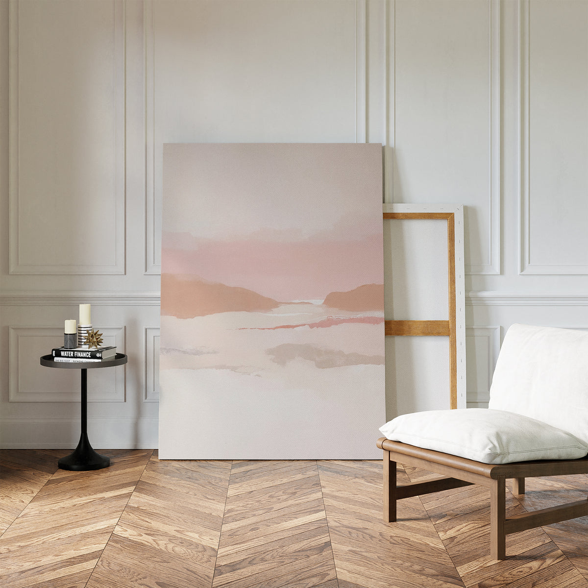 A minimalist room with herringbone wood floors, earth tones, a white cushioned chair, black side table with candles and books, and the Blush Mesa Canvas Art - Vertical leaning against a white paneled wall.